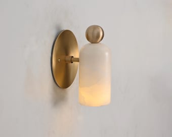 May include: A modern wall sconce with a brass finish and a cylindrical, off-white alabaster shade. The sconce features a round backplate and a small brass sphere on top of the shade. The light is warm and inviting.