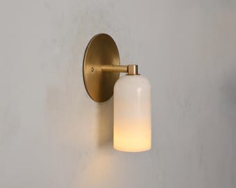 May include: A wall-mounted light fixture with a round, brass-colored backplate and a cylindrical, white glass shade. The shade emits a warm, diffused light. The fixture is attached to a brass-colored arm.