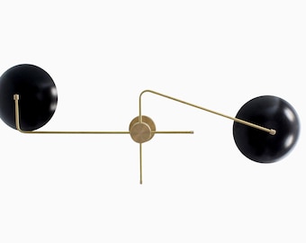 May include: A wall-mounted light fixture featuring two matte black spherical shades attached to thin, brushed brass arms. The central circular brass mount connects to a horizontal rod and an angled arm, supporting the two adjustable globes.