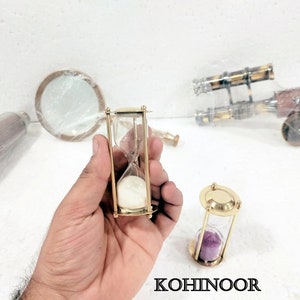 K&ouml;nnte beinhalten: Zwei Sanduhren aus Messing. Eine Sanduhr ist mit wei&szlig;em Sand gef&uuml;llt, die andere mit violettem Sand.