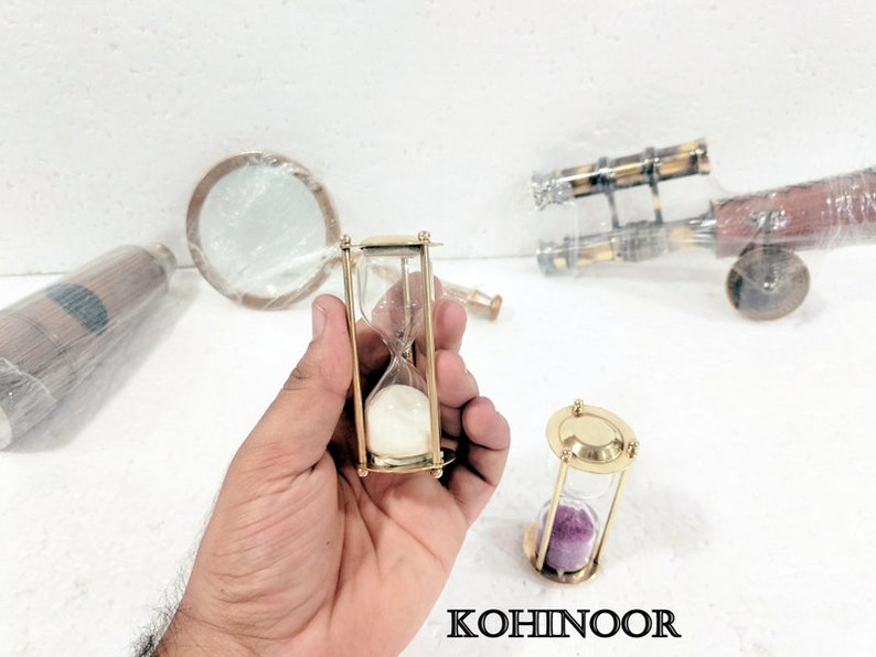 K&ouml;nnte beinhalten: Zwei Sanduhren aus Messing. Eine Sanduhr ist mit wei&szlig;em Sand gef&uuml;llt, die andere mit violettem Sand.