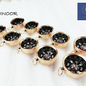 May include: A set of eight gold-toned compasses with black faces and white markings. The compasses are arranged in a semi-circle on a white surface. The compasses have a small loop at the top for attaching to a keychain or lanyard.