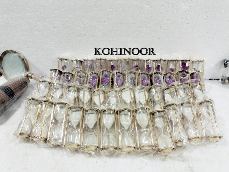 K&ouml;nnte beinhalten: Eine Gruppe von 25 goldgerahmten Sanduhren mit wei&szlig;em Sand. Die Sanduhren sind in durchsichtigem Plastik verpackt und in Reihen angeordnet. Das Wort "KOHINOOR" ist in schwarzen Buchstaben auf wei&szlig;em Hintergrund gedruckt.