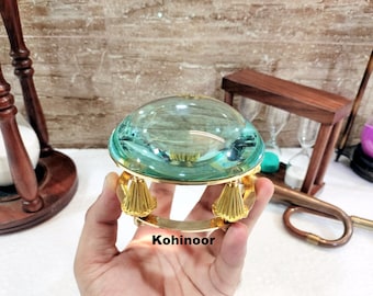 May include: A magnifying glass with a green glass lens and a gold metal base with three decorative feet. The base is inscribed with the word "Kohinoor".