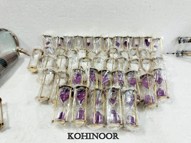 K&ouml;nnte beinhalten: Eine Gruppe von goldfarbenen Sanduhren mit lila Sand. Die Sanduhren sind in durchsichtigen Plastikt&uuml;ten verpackt und in Reihen angeordnet.