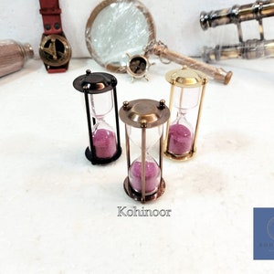 May include: Three hourglass sand timers with brass frames and pink sand. The hourglasses are on a white surface. The text "Kohinoor" is visible below the hourglasses.