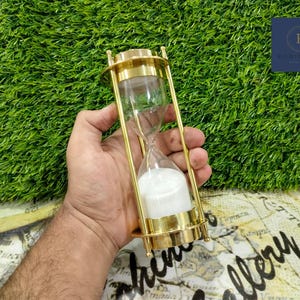 Peut inclure: Un sablier en or avec du sable blanc à l'intérieur. Le sablier est tenu dans la main d'une personne.