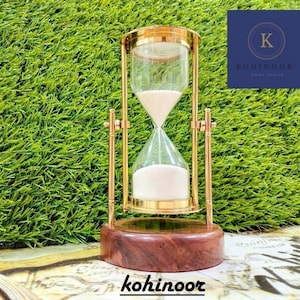 Può includere: Una clessidra in ottone con una base in legno. La clessidra è riempita di sabbia bianca e ha un bulbo in vetro trasparente. La base è incisa con la parola "kohinoor".