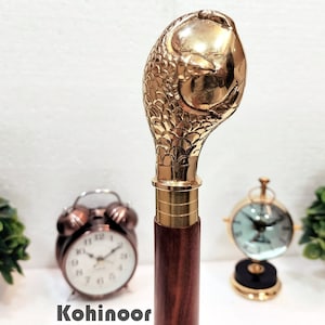 Op de afbeelding: Een gouden wandelstokgreep met een gedetailleerd ontwerp van een vis. De greep is bevestigd aan een bruine houten schacht.