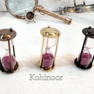 May include: Three hourglass sand timers with different coloured metal frames. The hourglasses are filled with pink sand and have a clear glass bulb. The frames are gold, brass, and black.