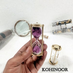 K&ouml;nnte beinhalten: Zwei Sanduhren aus Messing. Eine Sanduhr hat lila Sand und die andere wei&szlig;en Sand.