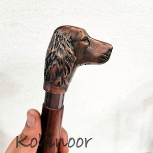 Peut inclure: Canne de marche en bois marron avec une tête de chien sculptée en détail. Le manche est brun foncé et la tête de chien est de couleur cuivrée. Le mot "Kohinoor" est visible en bas.