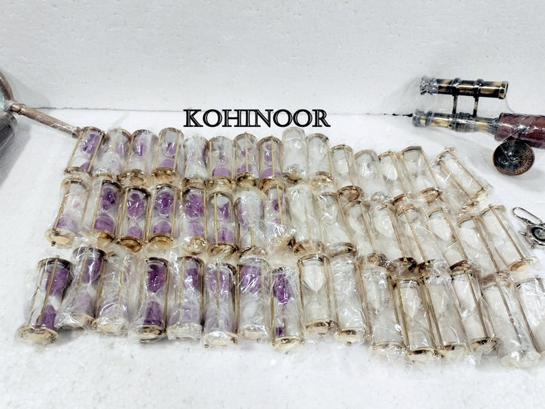 K&ouml;nnte beinhalten: Eine Gruppe von 30 kleinen, durchsichtigen Sanduhren-Anh&auml;ngern aus Glas mit goldfarbenen Metallrahmen und violettem Sand. Die Sanduhren sind in einzelnen Plastikt&uuml;ten verpackt.