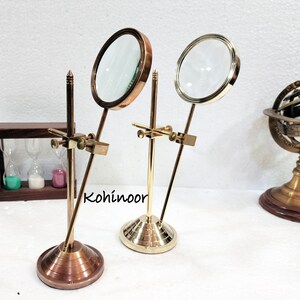 May include: Two brass magnifying glasses on stands. The magnifying glasses have round lenses and are mounted on adjustable arms. The stands have a round base and a vertical rod. The stands are made of brass and have a polished finish.