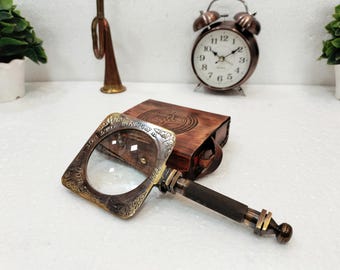 Puede incluir: Una lupa de estilo vintage con un marco cuadrado y ornamentado y un mango oscuro. La lupa está junto a una caja encuadernada en cuero y un reloj despertador de bronce. La estética general es antigua y decorativa.