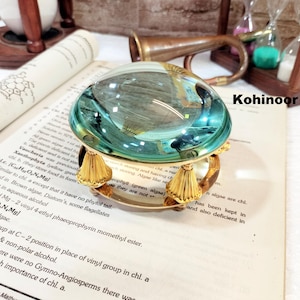 Op de afbeelding: Een vergulde loep met een ronde, heldere glazen lens en drie decoratieve gouden voetjes. De loep ligt op een boek met tekst over algen.