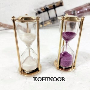 K&ouml;nnte beinhalten: Zwei Sanduhren aus Messing. Eine Sanduhr hat wei&szlig;en Sand, die andere violetten Sand. Die Sanduhren stehen auf einer wei&szlig;en Oberfl&auml;che. KOHINOOR ist auf der Oberfl&auml;che aufgedruckt.