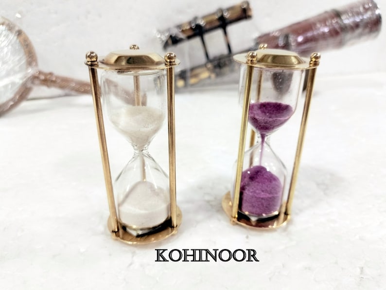 K&ouml;nnte beinhalten: Zwei Sanduhren aus Messing. Eine Sanduhr hat wei&szlig;en Sand, die andere violetten Sand. Die Sanduhren stehen auf einer wei&szlig;en Oberfl&auml;che. KOHINOOR ist auf der Oberfl&auml;che aufgedruckt.