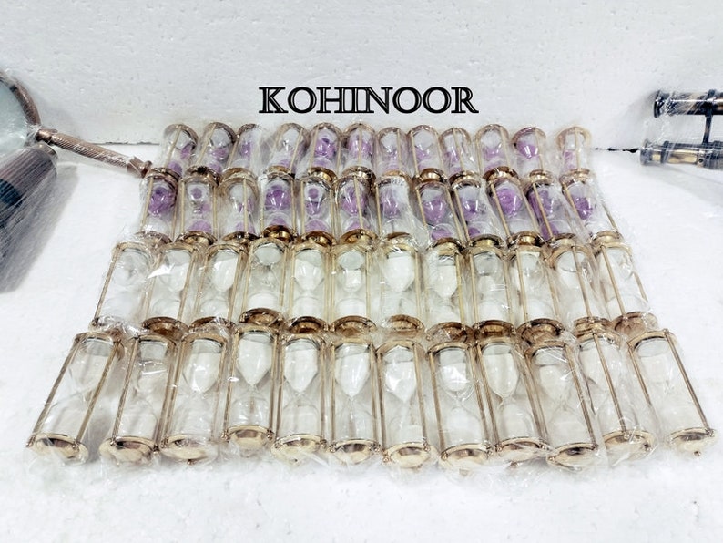 K&ouml;nnte beinhalten: Eine Gruppe von 40 goldfarbenen Sanduhren mit klaren Glaskolben. Die Sanduhren sind in Reihen auf einer wei&szlig;en Oberfl&auml;che angeordnet. Das Wort "KOHINOOR" ist in schwarzen Buchstaben auf wei&szlig;em Hintergrund &uuml;ber den Sanduhren gedruckt.