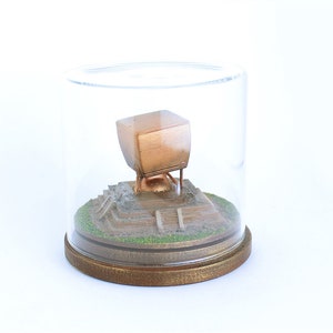 May include: A miniature replica of a building encased in a glass dome with a wooden base. The building is made of metal and has a square shape with a flat roof. The building is sitting on a small, dirt-covered platform with steps leading up to it.