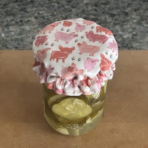 May include: A clear glass Mason jar filled with sliced pickles and dill, topped with a fabric cover featuring a repeating pattern of pink and coral-coloured pigs on a white background, secured with a ruffled edge around the jar lid.