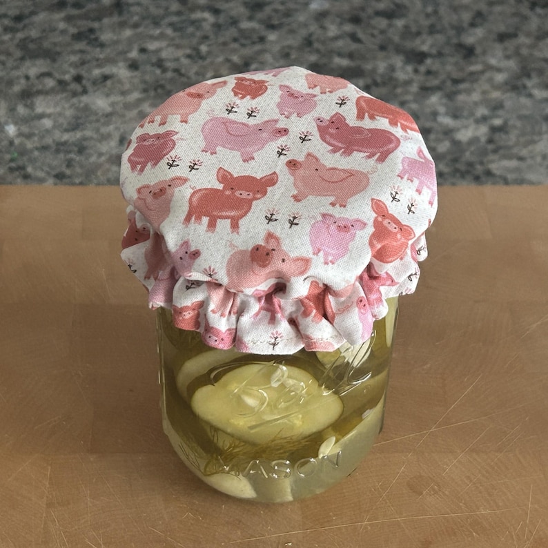 May include: A clear glass Mason jar filled with sliced pickles and dill, topped with a fabric cover featuring a repeating pattern of pink and coral-coloured pigs on a white background, secured with a ruffled edge around the jar lid.