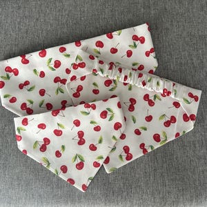 Könnte beinhalten: Ein Set aus Accessoires mit einem weißen Hintergrund und einem Muster aus roten Kirschen und grünen Blättern. Das Set enthält ein Stirnband und zwei dreieckige Bandanas.