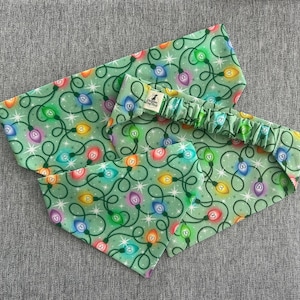 May include: A set of three pet accessories: a headband and two bandanas. The items feature a light green background with a repeating pattern of colorful Christmas lights and white stars. The headband has an elastic band.