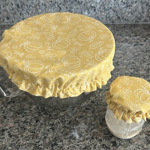 May include: Two round fabric bowl covers in a mustard yellow colour with a white line-art pattern of pumpkins, leaves, and acorns. The covers feature an elasticated, ruffled edge designed to fit securely over the rims of a large bowl and a small jar.
