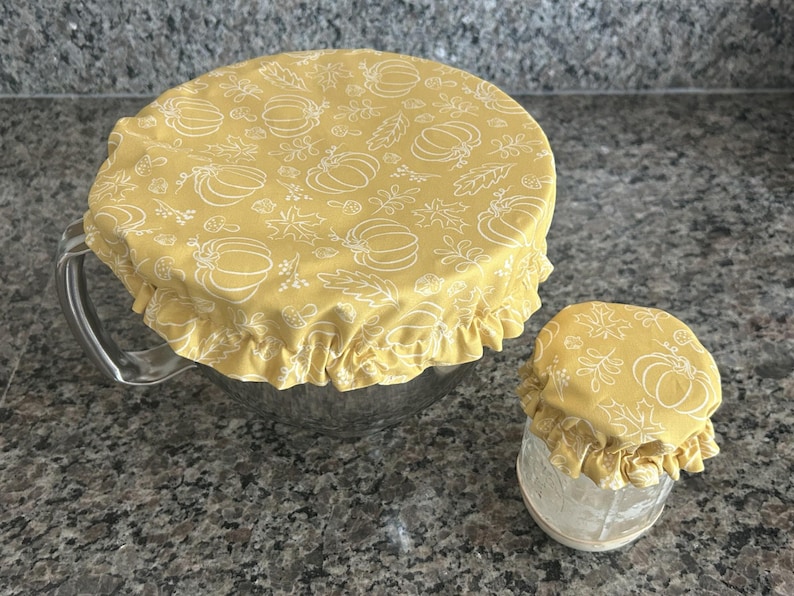 May include: Two round fabric bowl covers in a mustard yellow colour with a white line-art pattern of pumpkins, leaves, and acorns. The covers feature an elasticated, ruffled edge designed to fit securely over the rims of a large bowl and a small jar.