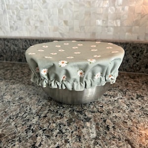 May include: A stainless steel bowl covered with a sage green fabric bowl cover featuring a repeating pattern of small white daisies with yellow centres. The fabric cover has an elasticated, ruffled edge that fits snugly around the bowl rim.