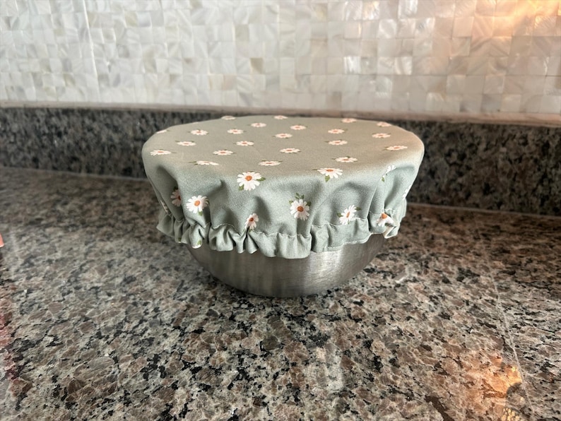 May include: A stainless steel bowl covered with a sage green fabric bowl cover featuring a repeating pattern of small white daisies with yellow centres. The fabric cover has an elasticated, ruffled edge that fits snugly around the bowl rim.