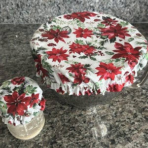May include: Two fabric bowl covers with elasticated edges, featuring a white background printed with red poinsettia flowers, green pine needles, and small brown pinecones. The covers are fitted over a large glass bowl and a small glass jar.