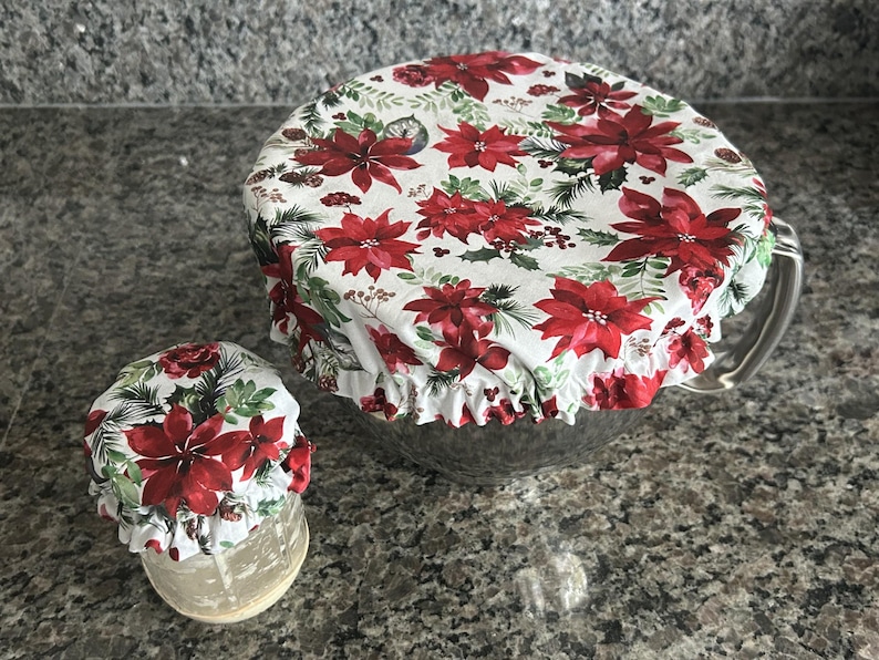May include: Two fabric bowl covers with elasticated edges, featuring a white background printed with red poinsettia flowers, green pine needles, and small brown pinecones. The covers are fitted over a large glass bowl and a small glass jar.