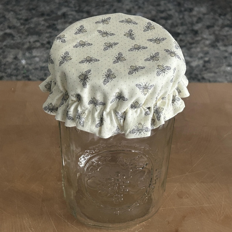 May include: A clear glass jar topped with a cream-coloured fabric cover featuring a repeating pattern of small grey bees. The fabric cover has a ruffled edge that hangs over the rim of the jar, which has a textured embossed design on its front surface.