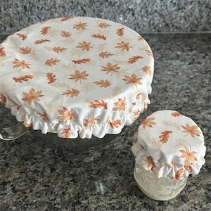 May include: Two round fabric bowl covers with an elasticated ruffled edge, featuring a white background printed with a repeating pattern of orange, brown, and yellow autumn leaves. One cover is large and fits a metal bowl, while the other is small.