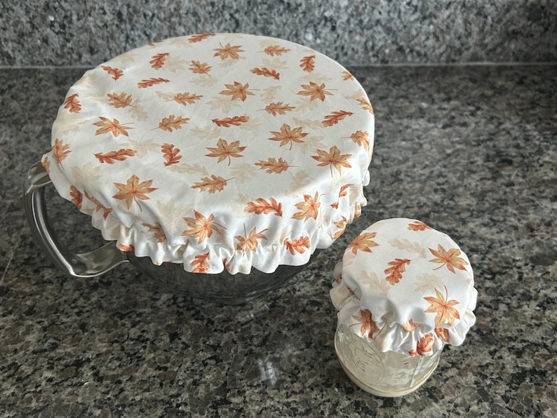 May include: Two round fabric bowl covers with an elasticated ruffled edge, featuring a white background printed with a repeating pattern of orange, brown, and yellow autumn leaves. One cover is large and fits a metal bowl, while the other is small.