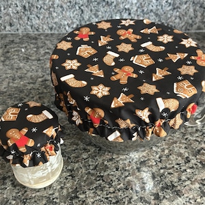 May include: Two elasticated fabric bowl covers featuring a black background with a repeating pattern of gingerbread people, Christmas stockings, gingerbread houses, and snowflakes. The covers are fitted over a small glass jar and a large glass bowl.