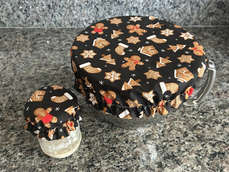 May include: Two elasticated fabric bowl covers featuring a black background with a repeating pattern of gingerbread people, Christmas stockings, gingerbread houses, and snowflakes. The covers are fitted over a small glass jar and a large glass bowl.
