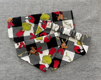 May include: Two pet bandanas and a matching headband. The bandanas and headband feature a black and white buffalo plaid pattern with cartoon Grinch and reindeer characters in red, green, and brown. The headband has a white and gray striped elastic.