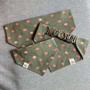 Puede incluir: Un conjunto de accesorios de tela verde oliva con un patrón repetido de calabazas naranjas y vides verdes. El conjunto incluye una diadema, un pañuelo para el cuello y un accesorio a juego. Se ve una pequeña etiqueta blanca con una ilustración negra.