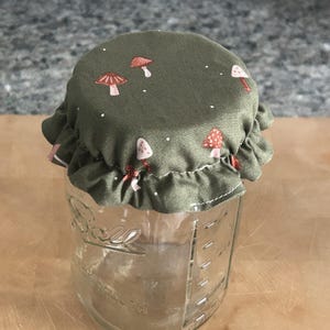 May include: A clear glass Ball brand mason jar topped with a fabric cover. The fabric is olive green and features a repeating pattern of small red and white mushrooms with tiny white dots. The cover has a ruffled edge and fits over the jar lid.