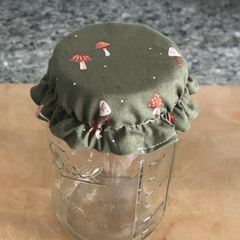 May include: A clear glass Ball brand mason jar topped with a fabric cover. The fabric is olive green and features a repeating pattern of small red and white mushrooms with tiny white dots. The cover has a ruffled edge and fits over the jar lid.