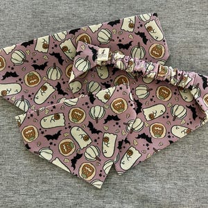 May include: A set of Halloween-themed accessories in a lavender color, featuring a headband, a bow, and a bandana. The fabric is patterned with white ghosts, pumpkins, bats, and cookies. The accessories are designed for pets.