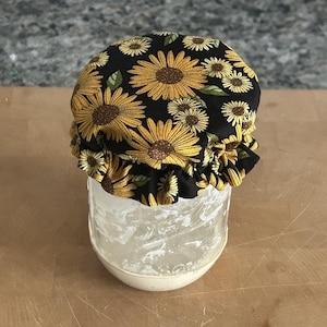 May include: A clear glass jar topped with a fabric cover featuring a black background and a repeating pattern of yellow sunflowers with green leaves. The fabric is gathered with a ruffled edge that fits snugly over the top of the glass container.