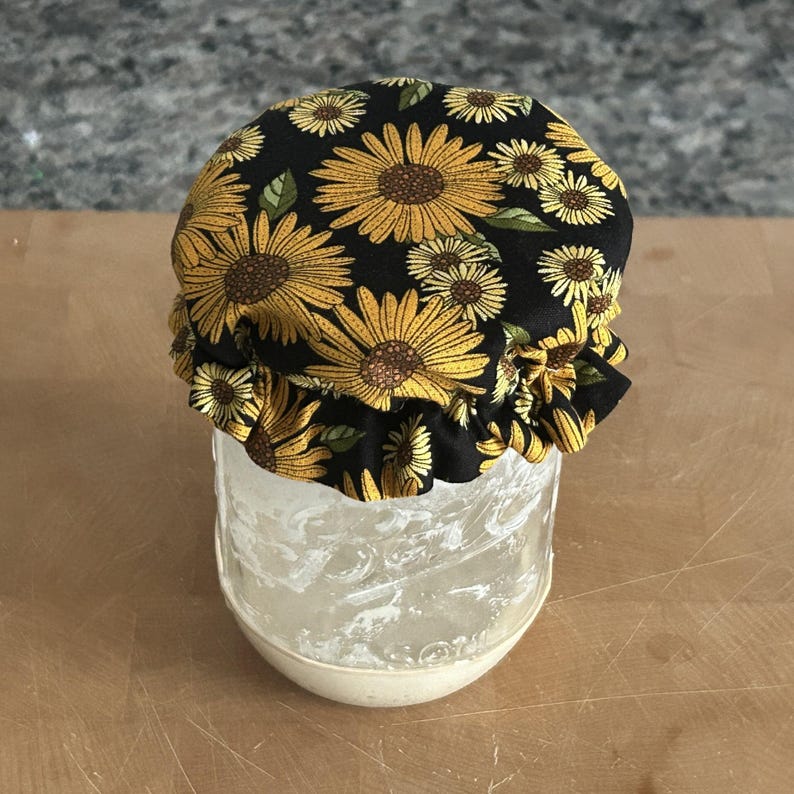 May include: A clear glass jar topped with a fabric cover featuring a black background and a repeating pattern of yellow sunflowers with green leaves. The fabric is gathered with a ruffled edge that fits snugly over the top of the glass container.