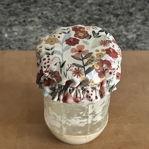 May include: A clear glass jar covered with a fabric lid topper featuring a floral pattern in shades of red, orange, yellow, and blue on a cream background. The fabric is gathered with an elastic band around the rim of the jar, creating a ruffled edge.