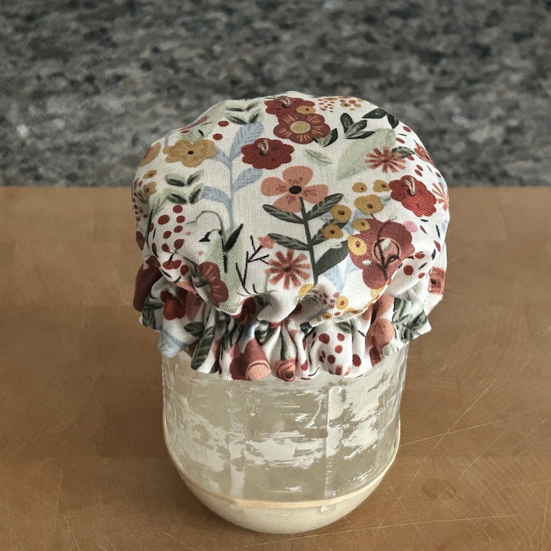 May include: A clear glass jar covered with a fabric lid topper featuring a floral pattern in shades of red, orange, yellow, and blue on a cream background. The fabric is gathered with an elastic band around the rim of the jar, creating a ruffled edge.
