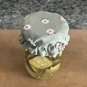 May include: A clear glass Ball mason jar filled with sliced pickles in brine, topped with a sage green fabric lid cover featuring a white and yellow daisy floral pattern and a ruffled edge that extends over the rim of the jar lid.