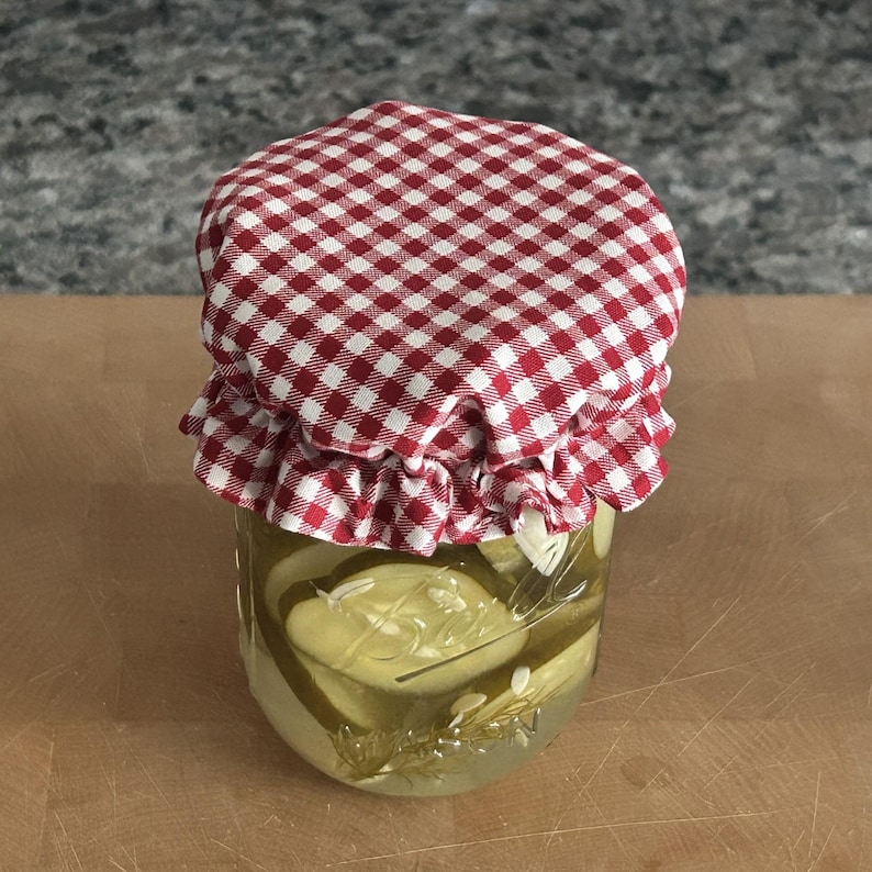 May include: A clear glass jar filled with sliced pickles in brine, topped with a red and white gingham fabric cover. The fabric is secured with a ruffled edge over the lid of the jar, which features embossed glass lettering on the side surface.